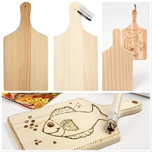 May include: Three wooden chopping boards with a handle. Two of the boards are plain and one has a fish design burned into the surface. The board with the fish design is being worked on with a wood burning tool.