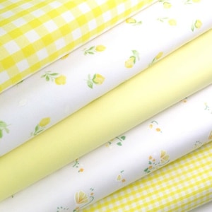 May include: Stack of four yellow and white fabric with different patterns. The top fabric is a yellow and white gingham pattern. The second fabric is white with a small yellow lemon print. The third fabric is a solid yellow. The bottom fabric is white with a small yellow floral print.