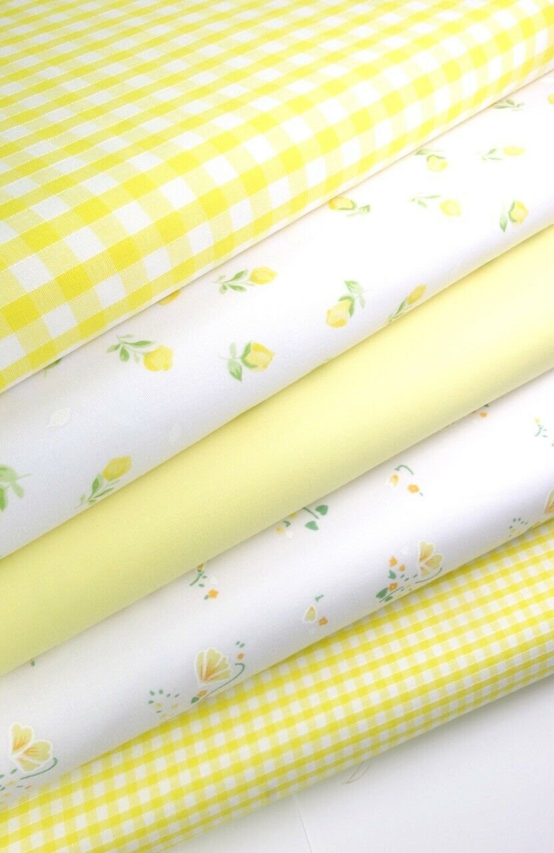 May include: Stack of four yellow and white fabric with different patterns. The top fabric is a yellow and white gingham pattern. The second fabric is white with a small yellow lemon print. The third fabric is a solid yellow. The bottom fabric is white with a small yellow floral print.