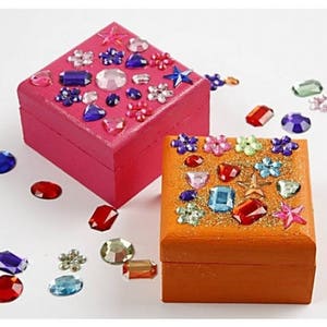 May include: Two small wooden boxes decorated with colorful jewels and glitter. The box on the left is pink and the box on the right is orange.