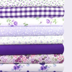 K&ouml;nnte beinhalten: Ein Stapel Stoffrollen in verschiedenen Blumen- und Vichy-Mustern. Die Stoffe weisen Farbt&ouml;ne von Lila, Lavendel und Wei&szlig; auf. Die Muster umfassen kleine Blumen, Rosen und ein klassisches Vichy-Karo. Die Rollen sind ordentlich gestapelt.