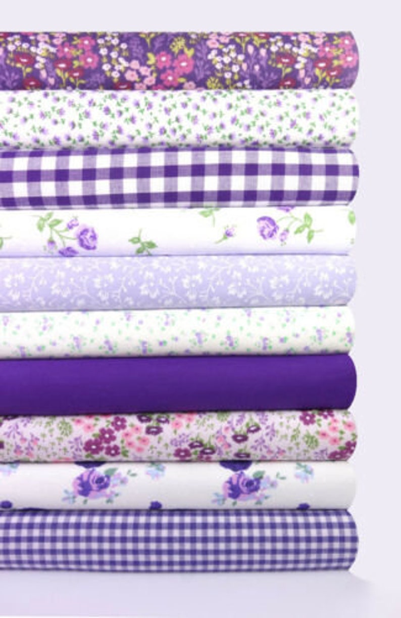 K&ouml;nnte beinhalten: Ein Stapel Stoffrollen in verschiedenen Blumen- und Vichy-Mustern. Die Stoffe weisen Farbt&ouml;ne von Lila, Lavendel und Wei&szlig; auf. Die Muster umfassen kleine Blumen, Rosen und ein klassisches Vichy-Karo. Die Rollen sind ordentlich gestapelt.