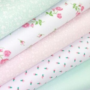 K&ouml;nnte beinhalten: Stapel von Stoffrollen in verschiedenen Pastellfarben. Die Stoffe weisen Blumen- und Punktmuster in Rosa, Wei&szlig; und Mintgr&uuml;n auf. Einige Rollen haben Rosen- und Blattdesigns, w&auml;hrend andere kleine Blumen- oder Punktmuster aufweisen.