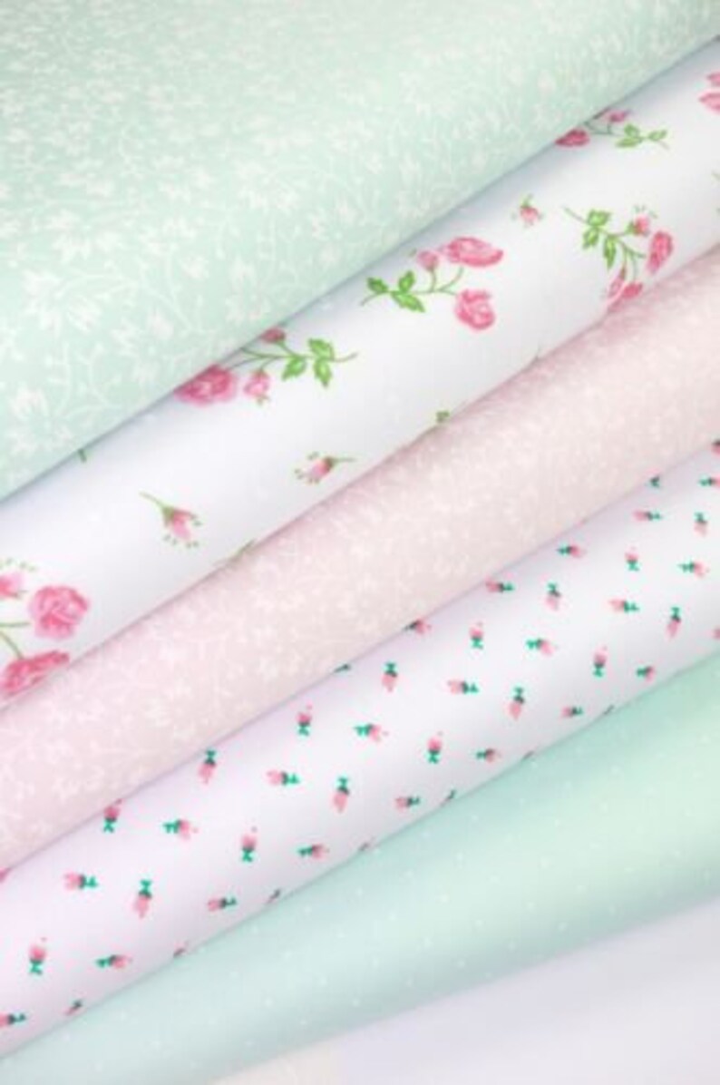 K&ouml;nnte beinhalten: Stapel von Stoffrollen in verschiedenen Pastellfarben. Die Stoffe weisen Blumen- und Punktmuster in Rosa, Wei&szlig; und Mintgr&uuml;n auf. Einige Rollen haben Rosen- und Blattdesigns, w&auml;hrend andere kleine Blumen- oder Punktmuster aufweisen.