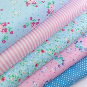 K&ouml;nnte beinhalten: Ein Stapel Stoffrollen mit verschiedenen Mustern. Die Stoffe zeigen Blumen-, Streifen- und Tupfenmuster in Blau- und Rosat&ouml;nen. Die Blumenmuster zeigen rosa Rosen und kleine Bl&uuml;ten.