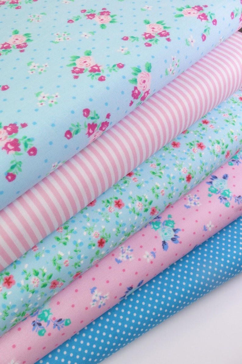 K&ouml;nnte beinhalten: Ein Stapel Stoffrollen mit verschiedenen Mustern. Die Stoffe zeigen Blumen-, Streifen- und Tupfenmuster in Blau- und Rosat&ouml;nen. Die Blumenmuster zeigen rosa Rosen und kleine Bl&uuml;ten.