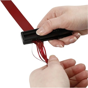 May include: A black ribbon cutter tool with a red ribbon being cut into multiple strands. The tool has a small window with a series of teeth that cut the ribbon.