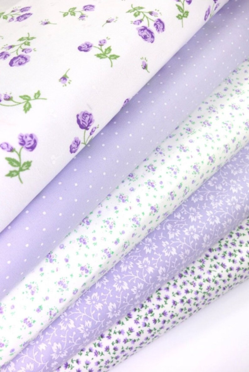 K&ouml;nnte beinhalten: Eine Auswahl an floralen Stoffrollen in Wei&szlig;- und Lavendelt&ouml;nen. Die Stoffe zeigen verschiedene Muster, darunter kleine lila Rosen, Tupfen und zarte Blumenranken. Ideal f&uuml;r Quilt- und Bastelprojekte.