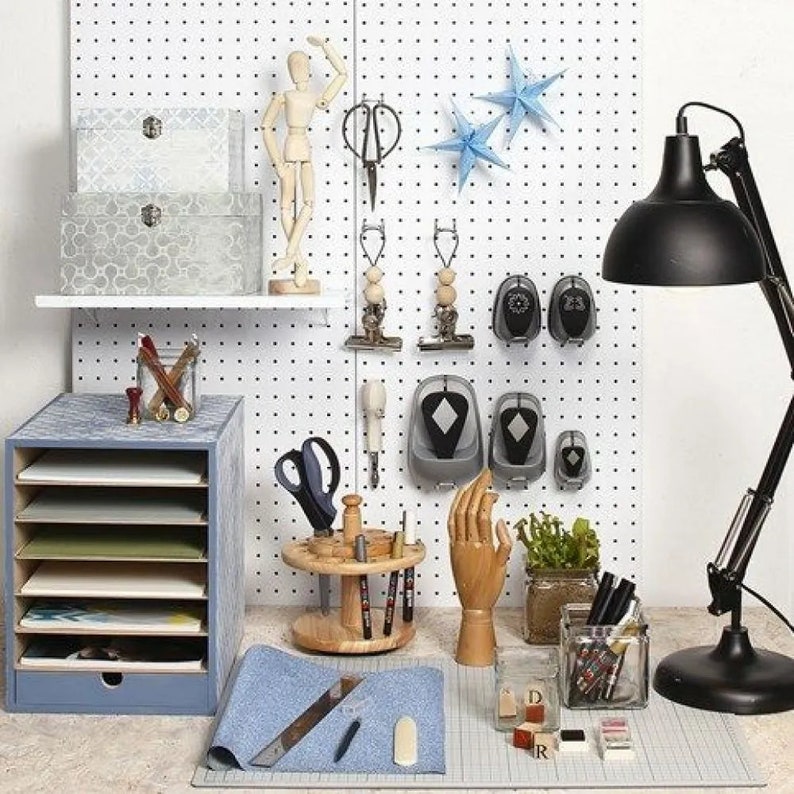 May include: A white pegboard with various tools and craft supplies, including a wooden mannequin, a set of blue star-shaped decorations, a wooden spool with paintbrushes, a set of hole punch tools, and a black desk lamp.