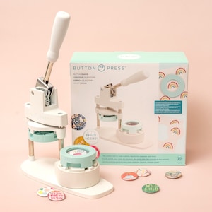 May include: A white button maker with a silver lever and a green base. The machine is on a pink surface with a box of buttons in the background. The box is light blue and green with rainbow designs and the text "Button Press".