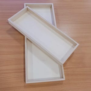 May include: Two rectangular wooden trays with light-coloured wood frames and off-white interiors are arranged on a wooden surface. One tray partially overlaps the other. The trays appear to be empty.