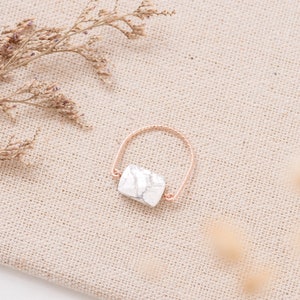 Peut inclure: Une bague en marbre blanc avec une bande en or rose. La bague est sur un fond en tissu beige.