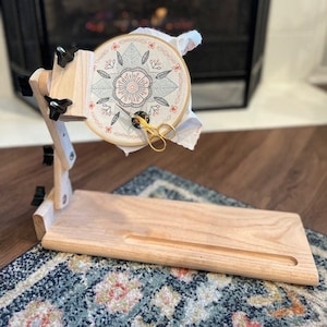 May include: A wooden embroidery hoop stand with a floral design embroidery hoop. The stand has a slot for holding a needle or scissors. The stand is on a patterned rug in front of a fireplace.