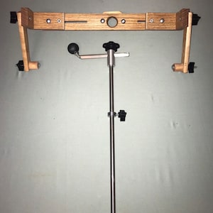 May include: A wooden frame with a metal rod and a crank handle. The frame has a hole in the center and is supported by two wooden arms. The rod is attached to the frame and has a knob at the top. The crank handle is attached to the rod and has a ball-shaped knob.