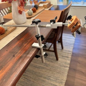 May include: A silver metal clamp with black knobs attached to a wooden table. The clamp has a long arm extending out with a smaller clamp at the end. The smaller clamp is holding a wooden block.