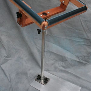 May include: A wooden embroidery hoop stand with a metal base and adjustable height. The stand has a black rubber roller on each side of the hoop.