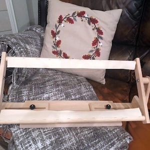 May include: A wooden embroidery hoop stand with a white fabric stretched across it. The stand has two adjustable arms and a base with a small tray. The stand is sitting on a brown leather chair with a gray blanket.