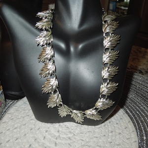 Puede incluir: Un collar plateado con un diseño de hojas. El collar está en un expositor negro.