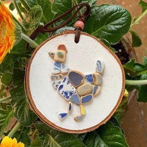 May include: A white wooden round ornament with a blue and white mosaic chicken design. The ornament has a brown leather cord with a bead.