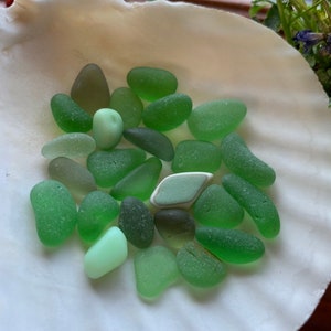 May include: A collection of smooth, sea glass stones in various shades of green, including a few lighter, almost white pieces. The stones are arranged on a white, textured surface.