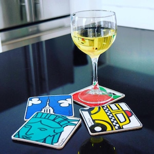 May include: A wine glass filled with a light yellow liquid sits atop a coaster with red, green, and white accents. Three additional coasters feature iconic New York City imagery: the Statue of Liberty, the Empire State Building, and a yellow taxi cab.
