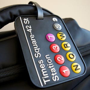 May include: Black luggage tag with white text that reads "Times Square-42 St Station". The tag has a loop for attaching to luggage and features colorful circular stickers with numbers and letters, including "N", "Q", "R", "W", "S", "7", "3", "2", and "1".