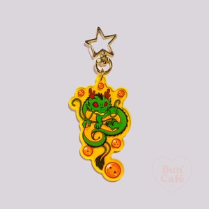 May include: A yellow acrylic keychain featuring a green dragon with red accents and orange spheres. The dragon is curled up on a yellow background. The keychain has a gold star-shaped clasp.