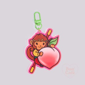 May include: A pink and red keychain featuring a cartoon monkey holding a peach. The monkey is wearing a red and gold outfit and a headband. The keychain has a green metal clip.