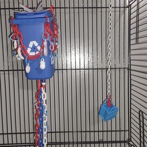May include: A blue plastic recycling bin toy with a white recycling symbol, adorned with red, white, and blue plastic chains and small charms. A separate vertical chain hangs nearby, featuring a small blue bucket charm and a red star-shaped bead.