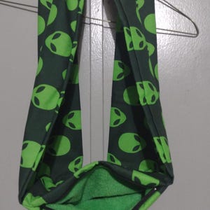 May include: A dark gray infinity scarf featuring a repeating pattern of bright lime green alien head silhouettes. The scarf has a soft, textured lime green interior lining and is draped over a wire hanger to display its circular loop shape.