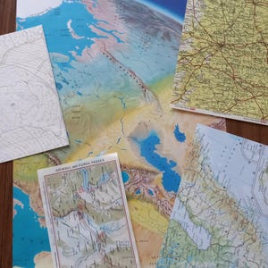 May include: A collection of overlapping paper maps featuring a large physical map of the Ural Mountains and Caspian Sea, a yellow road map, a topographic contour map, a map of the Grimsel and Furka Passes, and a coastal region map.