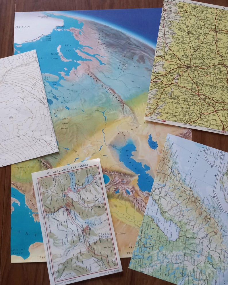 May include: A collection of overlapping paper maps featuring a large physical map of the Ural Mountains and Caspian Sea, a yellow road map, a topographic contour map, a map of the Grimsel and Furka Passes, and a coastal region map.