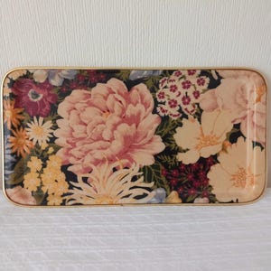 May include: A rectangular tray with rounded corners and a thin gold-toned metal rim. The surface features a dense floral pattern on a black background, displaying large pink peonies, cream-coloured blossoms, yellow clusters, and deep red flowers.