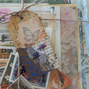 May include: A collection of assorted paper ephemera including vintage postage stamps, botanical prints, and map fragments, tied together with brown twine. A decorative tag features a butterfly illustration and a pressed blue flower on a textured background.