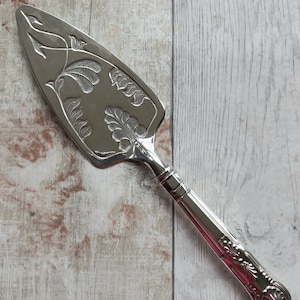 May include: Silver cake server with an ornate floral design. The handle is decorated with a scroll pattern.