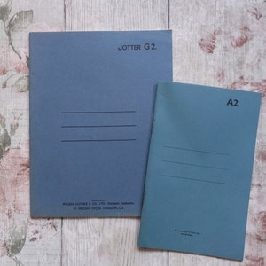 Peut inclure: Deux cahiers bleus, l'un intitulé "JOTTER G2." et l'autre "A2." Chacun comporte trois lignes horizontales. Les cahiers sont posés sur une surface à motifs floraux.
