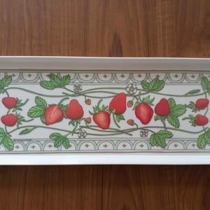 May include: A rectangular white ceramic serving tray featuring a central illustration of red strawberries, green leaves, and small white flowers. The design is framed by a decorative border with a repeating scalloped pattern in muted green and brown.