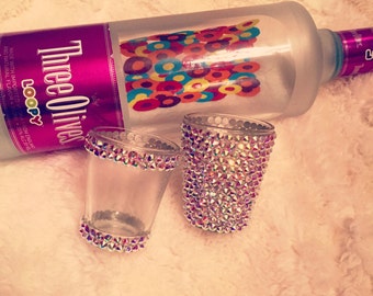 Op de afbeelding: Een matglazen fles Three Olives Loopy wodka met twee shotglaasjes. Eén shotglaasje is bedekt met iriserende strass-steentjes, en de andere heeft strass-steentjes rond de rand en de basis. De fles heeft een kleurrijk etiket.