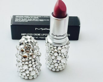 Op de afbeelding: Een zilveren lipstickbuis met een roze lipstickkogel. De buis is bedekt met heldere strasssteentjes. De tekst "AMPLIFIED CREME LIPSTICK" is zichtbaar op de zwarte doos.
