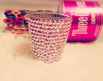 Op de afbeelding: Een helder shotglaasje bezet met iriserende strass-steentjes. Op de achtergrond staat een roze blikje Three Olives wodka. Er zijn ook kleurrijke, cirkelvormige decoraties.