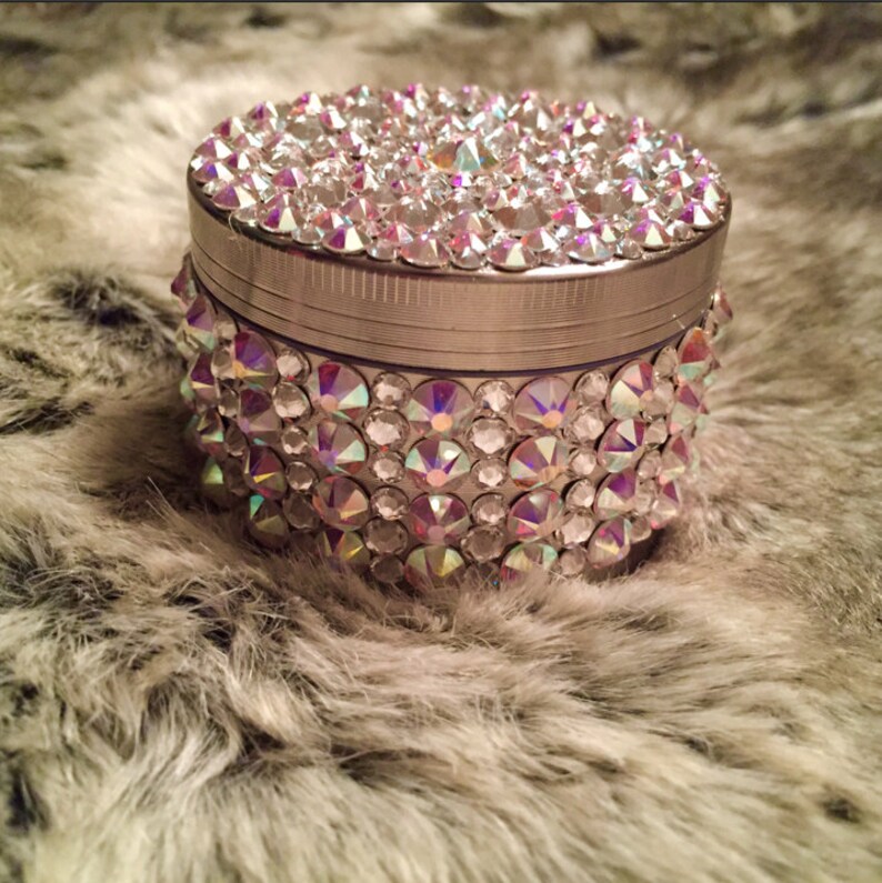 Op de afbeelding: Een zilveren grinder met een rond, helder kristal-achtig ontwerp. De grinder is bedekt met kleine, iriserende strass steentjes.