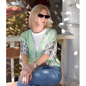 May include: A light green open-front cardigan with a geometric pattern on the sleeves, worn over a white t-shirt and blue jeans. The cardigan has a floral design and is paired with sunglasses and a watch.