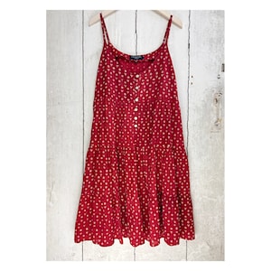 Puede incluir: Un vestido rojo sin mangas con estampado floral blanco y botones. El vestido tiene una falda de capas y está hecho de una tela ligera.