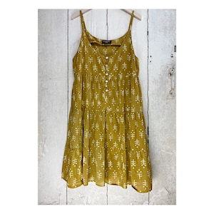 Puede incluir: Un vestido amarillo mostaza de capas con un patrón geométrico blanco. El vestido tiene tirantes finos y un frente abotonado.