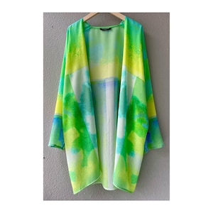 May include: A lightweight, open-front kimono in a vibrant watercolor design. The garment features a blend of green, yellow, blue, and white hues. The kimono is hanging on a wooden hanger against a neutral-coloured wall.