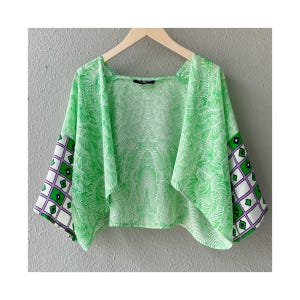 May include: A green and white patterned kimono-style top. The body of the top features a repeating leaf design. The sleeves have a geometric pattern in green, white, and purple. The top is hanging on a wooden hanger.