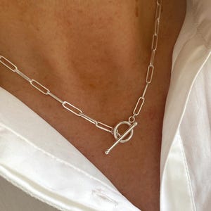 May include: A silver chain necklace with an elongated oval link design. The necklace features a toggle clasp closure. The necklace is worn over a white button-down shirt.