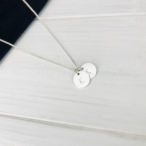 May include: A silver-coloured chain necklace featuring two small, circular, flat pendants. Each pendant is engraved with a single uppercase letter, specifically the letters K and L, suspended from a delicate, thin, metallic link chain.