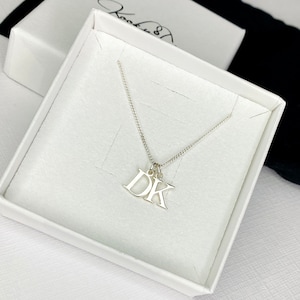 May include: A silver necklace with a pendant featuring the initials "DK" in a simple, modern font. The necklace is displayed in a white box.