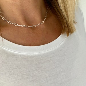 May include: A silver chain necklace with a delicate, oval-shaped link design. The necklace is worn by a person with a white shirt.